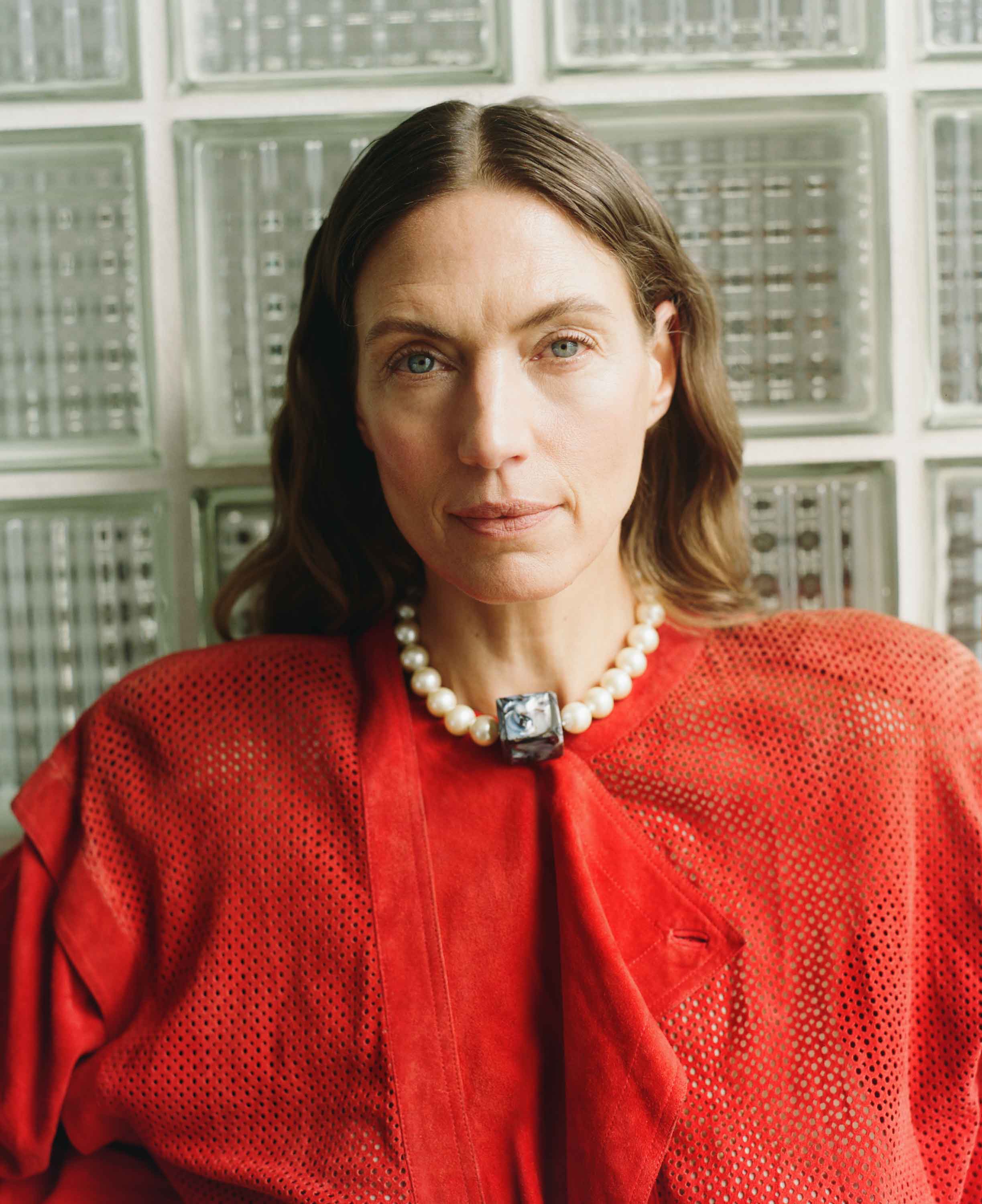 Woman wearing a red outfit with a pearl necklace against a glass block wall.