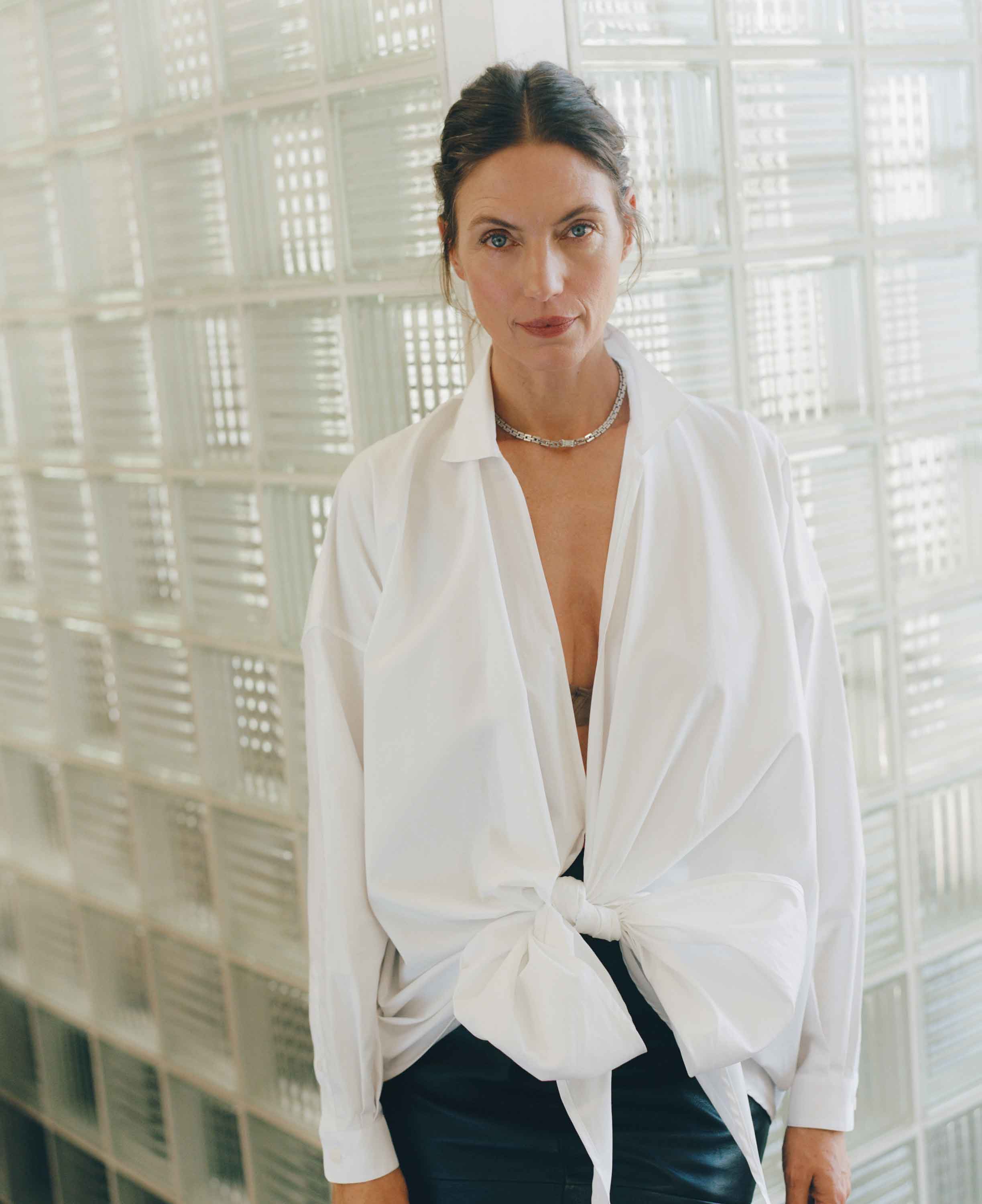 Woman wearing a white blouse with a knot detail in front of a glass brick wall.